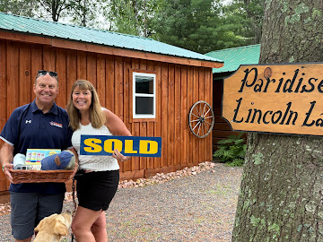 Two clients outside a wood-sided cabin, holding a SOLD sign